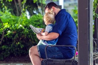 A child sitting on a man's lap being read a story outside on a bench.