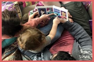 A child sitting on an adult's lap, pointing to a story, with another adult beside them.