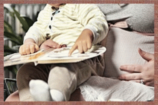A child pointing at a page of a book while sitting on his pregnant mother's lap.
